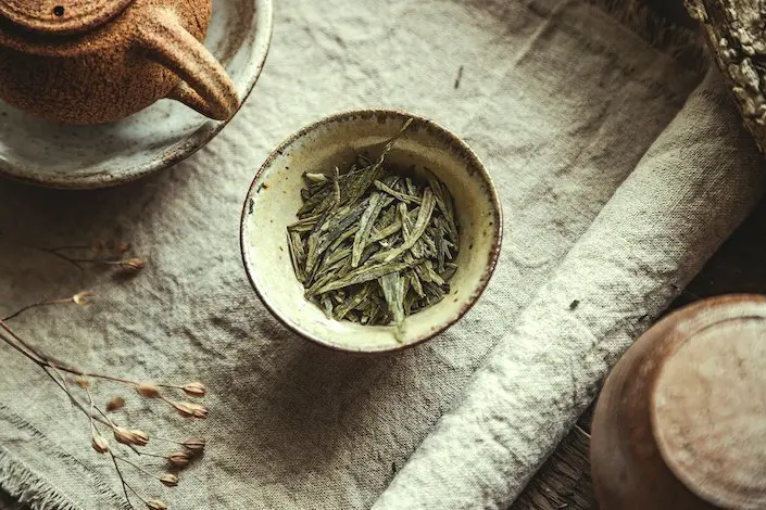 a Chinese tea cup filled with flat green tea leaves and a partially visible tea pot sitting on a table