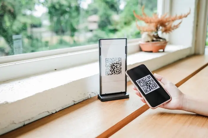 a hand holding a phone displaying a QR code next to another phone with QR code sitting straight up on a wooden table